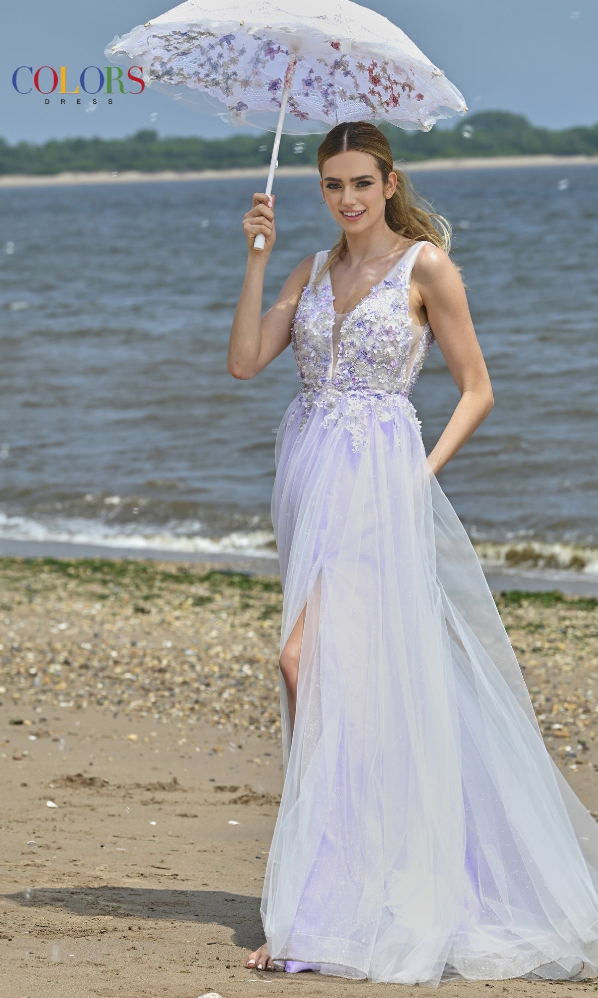 Wearing the Glitter-Tulle Long Prom Ball Gown: Colors Dress 3499, a woman in a light purple, floral-embellished gown holds a white parasol on a sandy beach near the water.